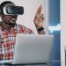 A man wearing VR glasses working on laptop, immersed in augmented virtual reality, sitting at office desk. Black young businessman using innovation technology for work and business.